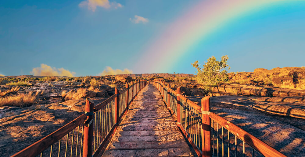 A path through the desert with fencing on either side. A rainbow shines in the distance.
