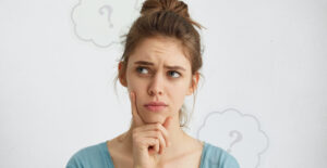 Young woman with a questioning look on her face and her hand on her chin. Low opacity thought bubbles with question marks hover in the background.