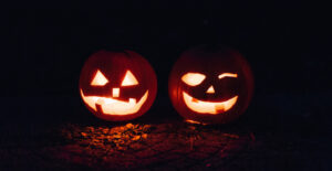 Light glows through the carved faces of a pair of jack o'lanterns sitting on a dimly lit ground.