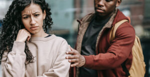 A young couple who are upset. The woman faces the camera, her face uphappy. The man stand behind her, reaching forward to touch her shoulder.