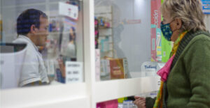 A woman in a sweater and wearing a face mask approaches a pharmacist working behind a window.