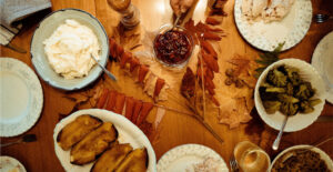 Overhead view of a Thanksgiving table decorated with leaves and laden with mashed potatoes, roasted squash, stuffing, broccoli, and cranberry sauce. One plate is already filled with sliced turkey, and a hand reaches for the cranberry sauce.
