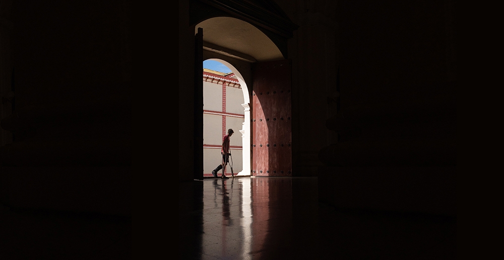 In a large, dark space, a young man using crutches passes a brightly lit doorway.