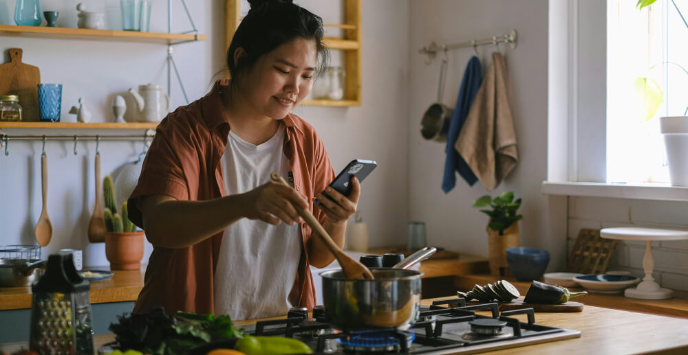 A young woman consults her phone as she stirs a saucepan