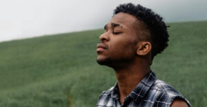 A young African American man stands in a grassy field, with his eyes closed and a pensive expression on his face.