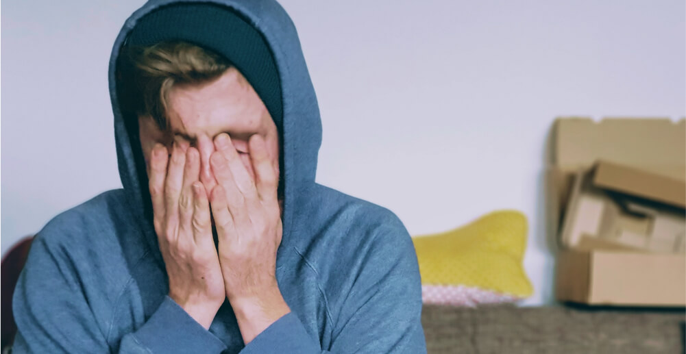 Young white man in a gray hoodie rubs his face in distress.