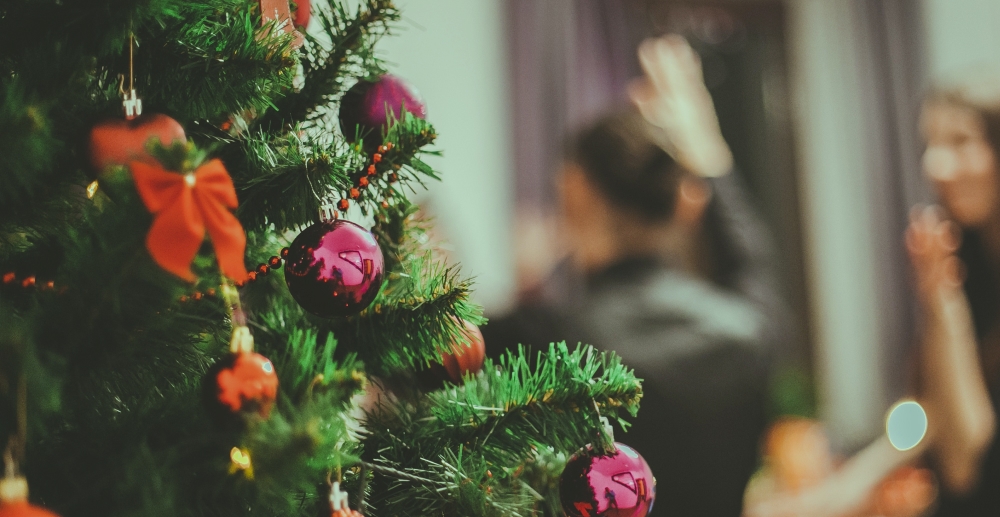 A decorated Christmas tree is in the foreground, and people are out of focus in the background.