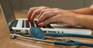 Close-up on a woman's hands as she types on a laptop. A stethoscope lies on the desk beside the computer.