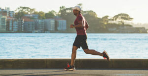 A man in a tee shirt, shorts, and a baseball cap jogs in front of a body of water.