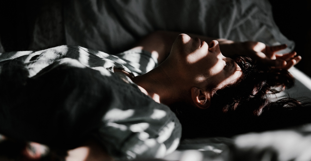 A person lies on a bed, with bars of light from the window blind across their face