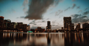 The skyline of downtown Orlando, Florida at dusk, with Lake Eola in the foreground and tall buildings behind it.