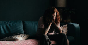 A person with long red hair sits on a sofa in a dark room with their head in their hands