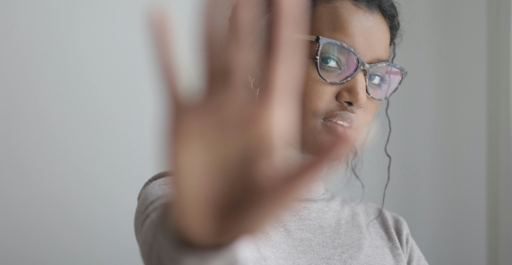 A young, Black woman in glasses holds. a hand up in a Stop" gesture, blocking the view of the camera