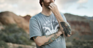 A man with tattoos on his arms and a Volcom t-shirt stands in front of a background of desert mountains. One hand is in front of his chin in a thoughtful pose.