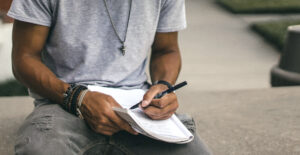 A man sits on a bench outdoors, writing in a notebook,