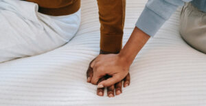 Focus on the hands of a couple sitting on a bed. The man's hand is pressed flat against the bed, and the woman's hand is on top of it, holding it.