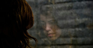 Reflection in the window of a sad-faced White woman with long hair. The view beyond her reflection is of a brick wall