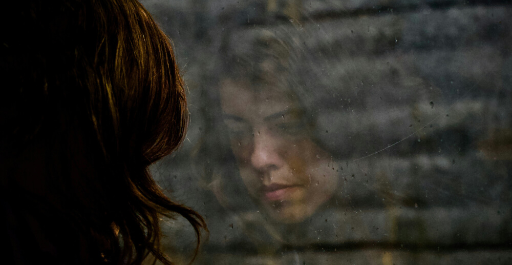 Reflection in the window of a sad-faced White woman with long hair. The view beyond her reflection is of a brick wall