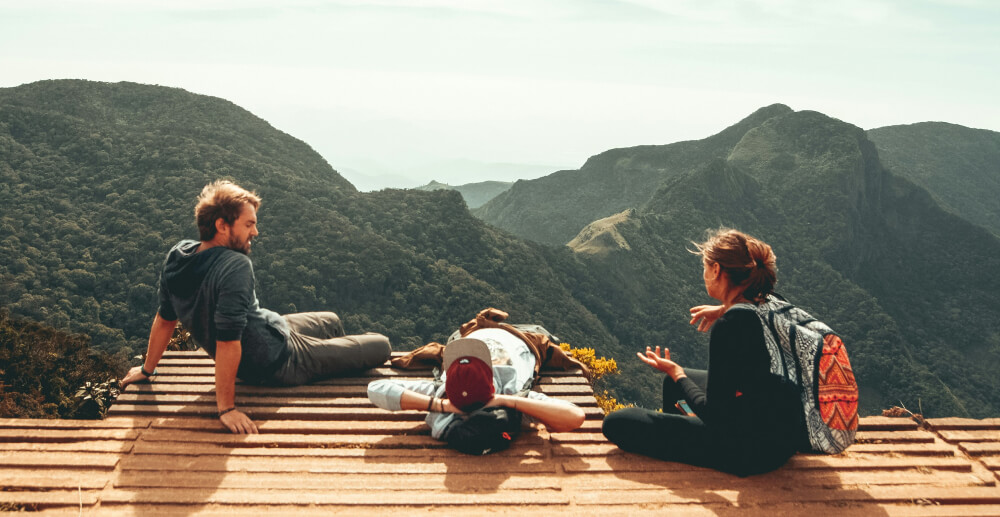 View from behind of three people sitting on a platform at the top of a mountain, with a view of other green peaks.