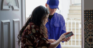 In an open doorway, a woman signs for a delivery on a screen held out to her by a man in a baseball cap.