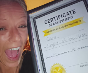 Gloria smiles wide, holding up a certificate of achievement