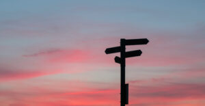 A street sign pointing in multiple directions is silhouetted against a pink and blue sunset