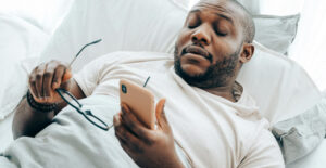 A Black man with a trimmed beard lies against white pillows, looking at his smartphone. In one hand, he holds a pair of glasses.