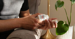 A person leans over the arm of a couch, shaking a generic buprenorphine/naloxone tablet into their hand. The image is a closeup on the hands that does not show the person's face.