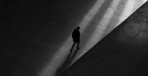 Black and white photo from above of a lone person crossing a wide, concrete space. The photo is moodily lit.