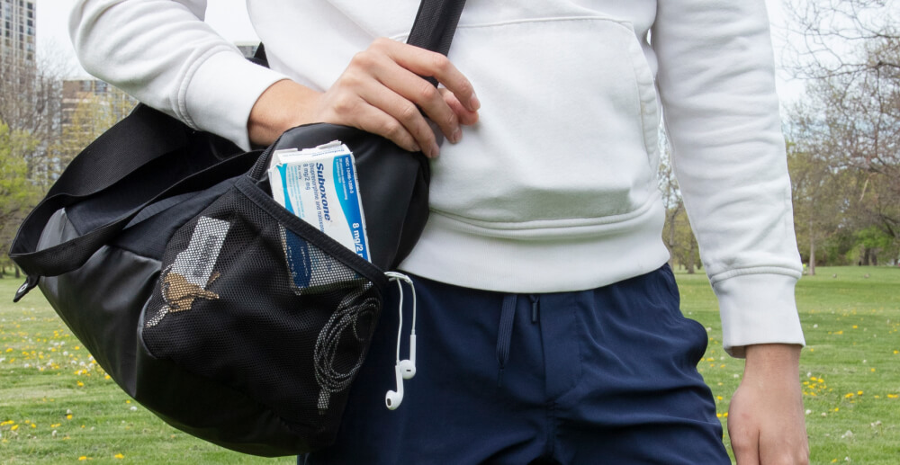 A man in a sweatshirt stands outdoors, carrying a gym bag. Visible through the mesh pocket of the bag is a box of Suboxone.