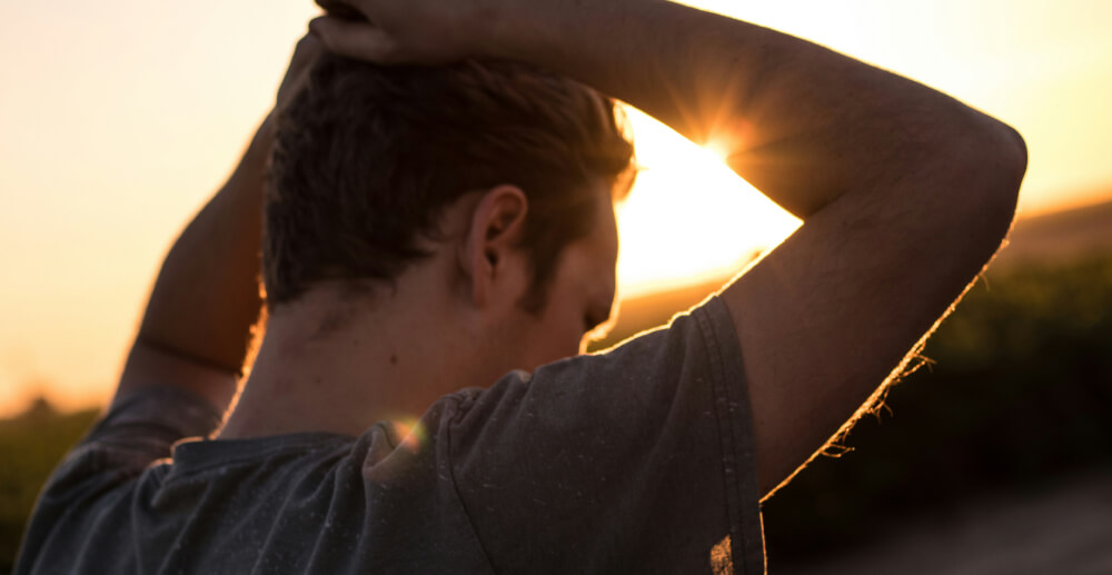 A yound man stands outdoors with his hands on his head in a resigned manner. The sunset is bright behind him.