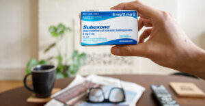 A hand holds a slightly battered box of Suboxone up to the camera. Beyond the box is a table holding a newspaper, a pair of glasses, and a mug.