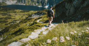 A man walks away from the camera along a winding trail on a lush mountainside.