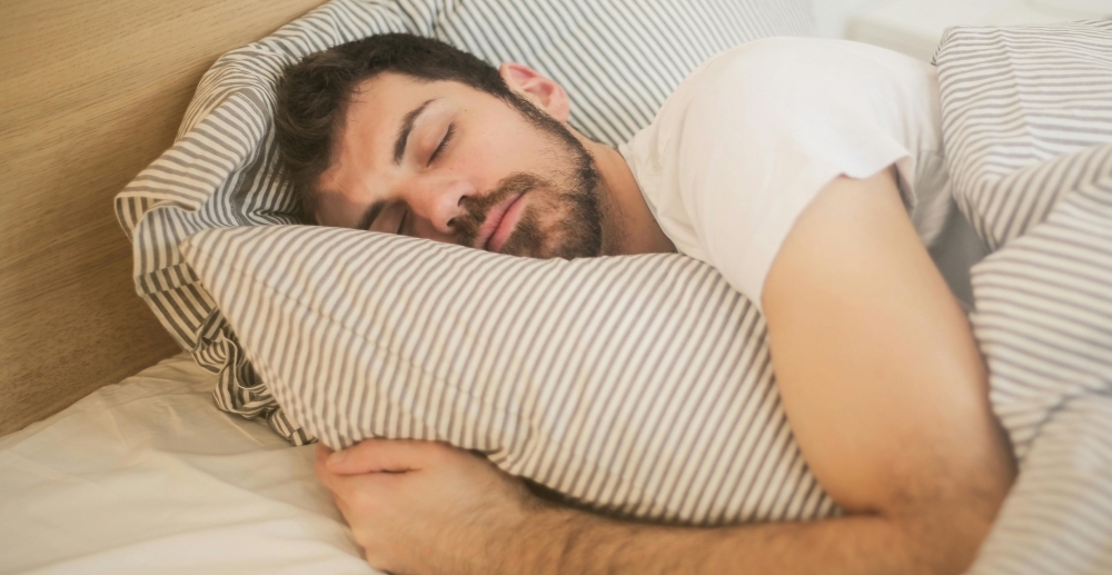 A man with a short, brown beard hugs his pillow to him as he sleeps.