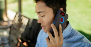 A young, Asian man sits on a bench outdoors, talking on a cell phone