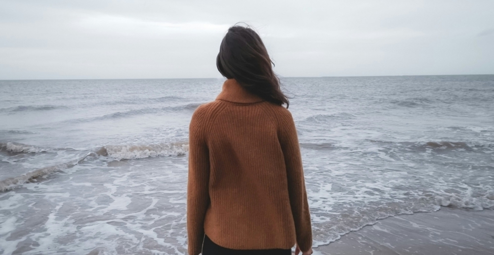 View of the back of a woman in a brown sweater as she stares out at the ocean.