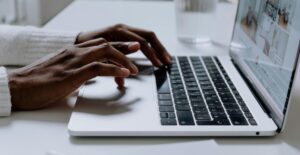 Dark-skinned hands type on a laptop on a clean, white desk.