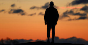 A man stands along on a hilltop, silhouetted by the red of the setting sun