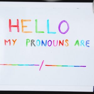 A person holds a small, dry-erase board with text written in rainbow markers "Hello, my pronouns are _/_". A Journey to Self-Love