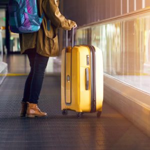 A woman with a yellow rolling suitcase and a blue and purple backpack stands on the moving walkway in an airport. Vacation sober, traveling sober.