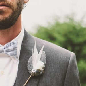 Closeup of a groom on his wedding day, wearing a gray suit and a white tie. He has a boutonierre made of feathers.