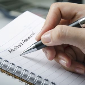 Closeup of a notebook, with a hand holding a pen to the page. The words, "Medicaid Renewal" are written.