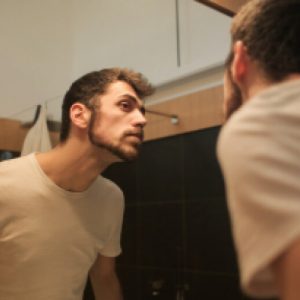 A young man in a white t-shirt looks at himself in the mirror