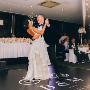 A bride and groom laugh as they dance alone at their wedding. Guests watch from their reception tables, and sparklers burn near the edge of the dance floor.