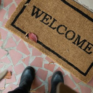 A welcome mat in front of the door to a house. The point of view looks down at the viewer's feet in front of the welcome mat.