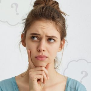 Young woman with a questioning look on her face and her hand on her chin. Low opacity thought bubbles with question marks hover in the background.