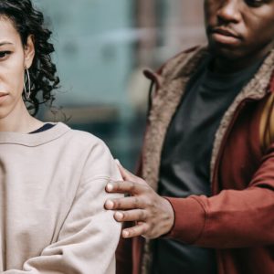 A young couple who are upset. The woman faces the camera, her face uphappy. The man stand behind her, reaching forward to touch her shoulder.