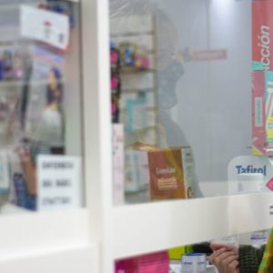 A woman in a sweater and wearing a face mask approaches a pharmacist working behind a window.