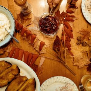 Overhead view of a Thanksgiving table decorated with leaves and laden with mashed potatoes, roasted squash, stuffing, broccoli, and cranberry sauce. One plate is already filled with sliced turkey, and a hand reaches for the cranberry sauce.