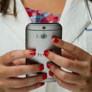 A female doctor in a white coat with a stethoscope around her neck uses a smartphone. The composition of the shot focuses on her hands, with her face out of frame.